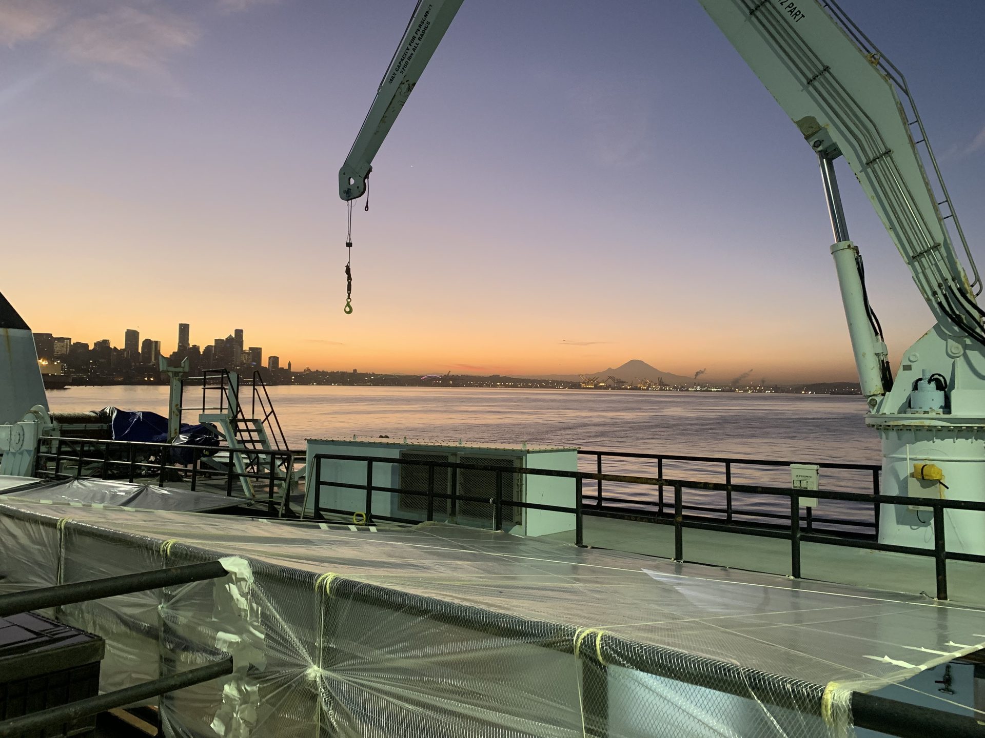 Deck of a working vessel at sunrise, with deck cranes overhead and a city skyline and snow-capped mountain across the water